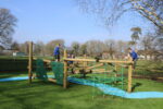 Children in school uniforms play on a wooden climbing structure resembling Pick Up Sticks in an outdoor playground under a clear sky.