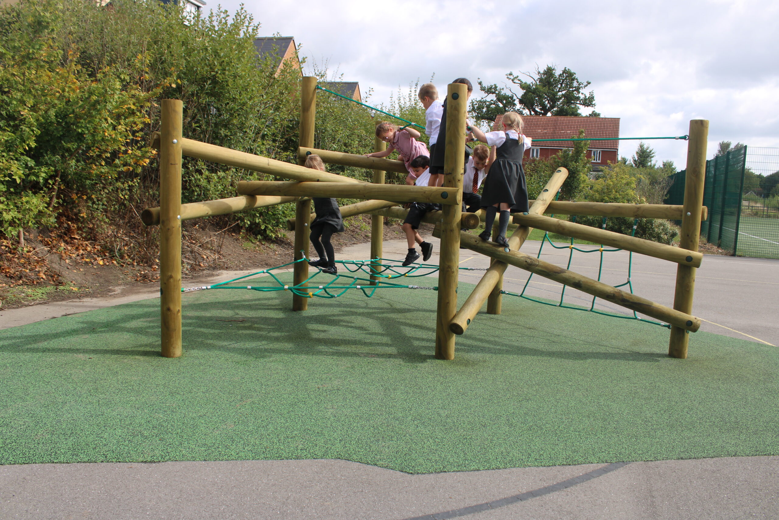 Several children play on a wooden climbing frame with rope sections, reminiscent of a giant Pick Up Sticks game, in an outdoor playground bordered by greenery and pavement.