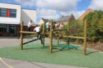 Five children in school uniforms climb on a wooden and rope play structure, reminiscent of Pick-up Sticks, in a school playground on a sunny day.