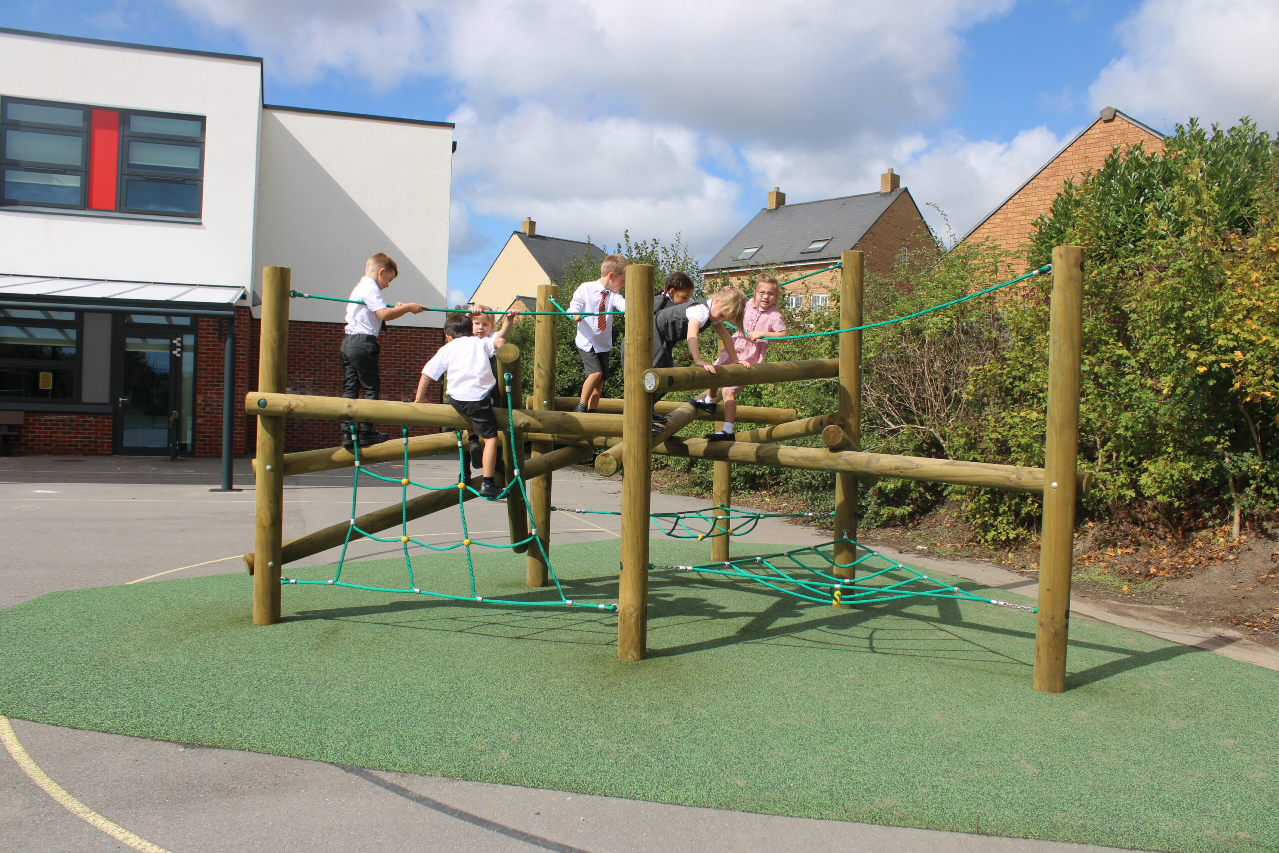 Five children in school uniforms climb on a wooden and rope play structure, reminiscent of Pick-up Sticks, in a school playground on a sunny day.