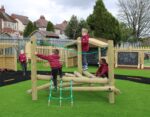 Four young children in maroon uniforms climb and play on a wooden playground structure in a fenced outdoor area with artificial grass, reminiscent of a lively game of Pick-up Sticks.