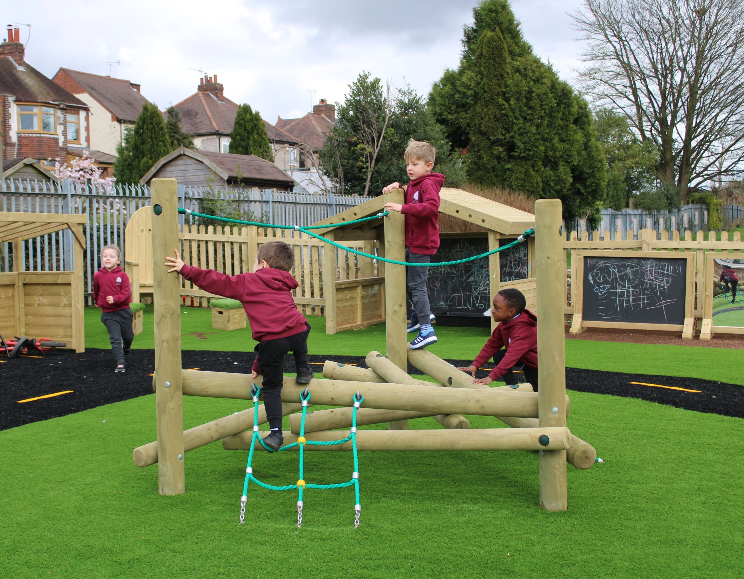 Four young children in maroon uniforms climb and play on a wooden playground structure in a fenced outdoor area with artificial grass, reminiscent of a lively game of Pick-up Sticks.