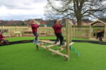 Children play and climb on wooden equipment in an outdoor playground with artificial grass and fencing, reminiscent of a giant Pick Up Sticks game, under a cloudy sky.