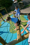 Three children in school uniforms play on a rope and log climbing structure, enjoying the playground’s Pick Up Sticks (4) with Poly Slide on a sunny day.