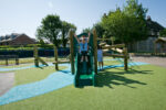 Two children play on a Pick Up Sticks (4) with Poly Slide, enjoying the sunny outdoor playground surrounded by trees and houses.