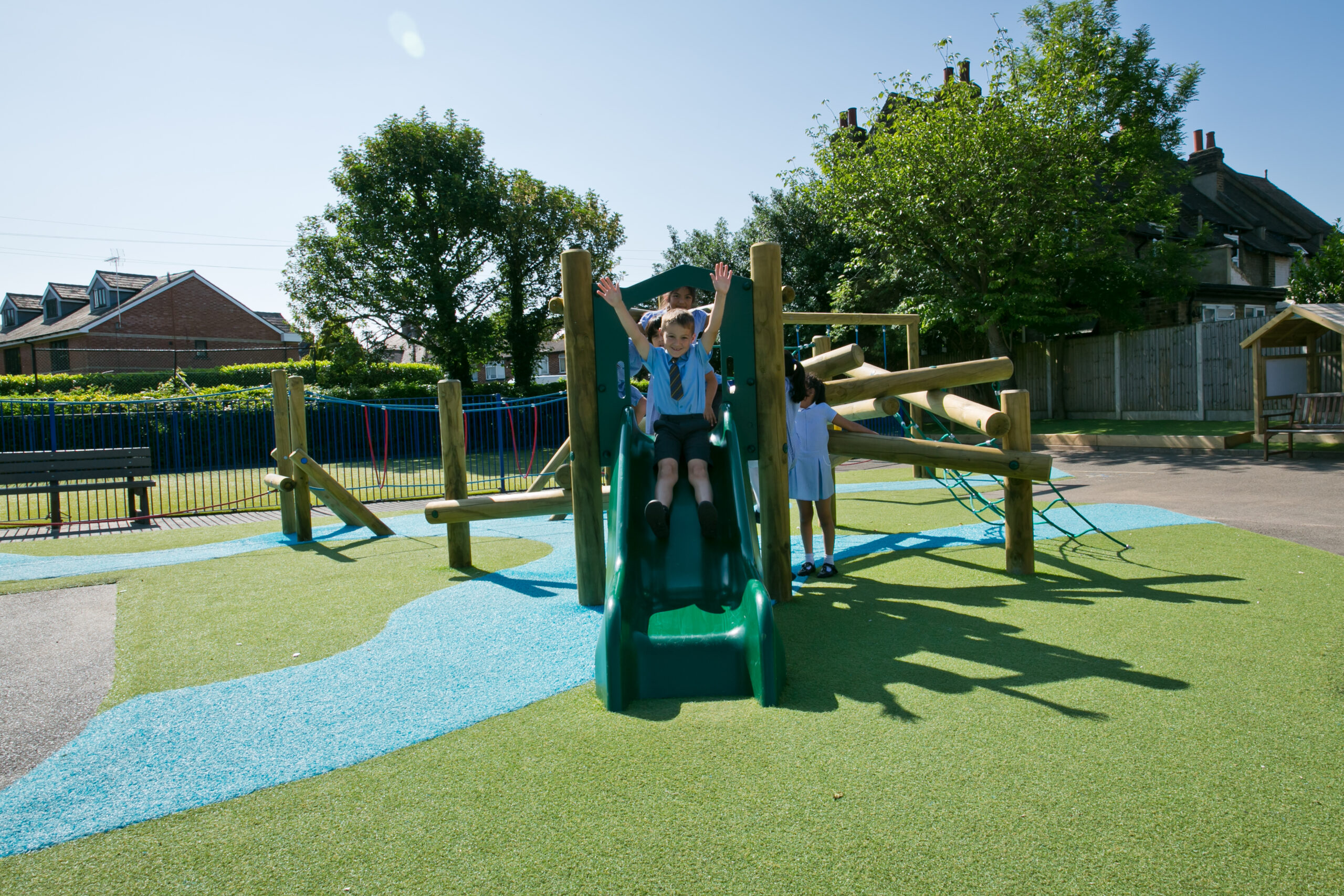 Two children play on a Pick Up Sticks (4) with Poly Slide, enjoying the sunny outdoor playground surrounded by trees and houses.