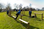 Children in school uniforms play and climb on wooden balance beams and structures in an open grassy field on a sunny day.