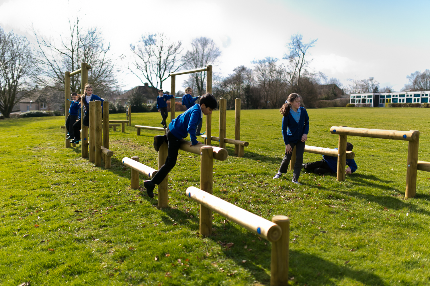 Children in school uniforms play and climb on wooden balance beams and structures in an open grassy field on a sunny day.