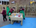 Three young children in green aprons play with water and plastic toys at a wooden sensory table outdoors, near a wooden fence, enjoying one of the 7 Benefits of Outdoor Play.