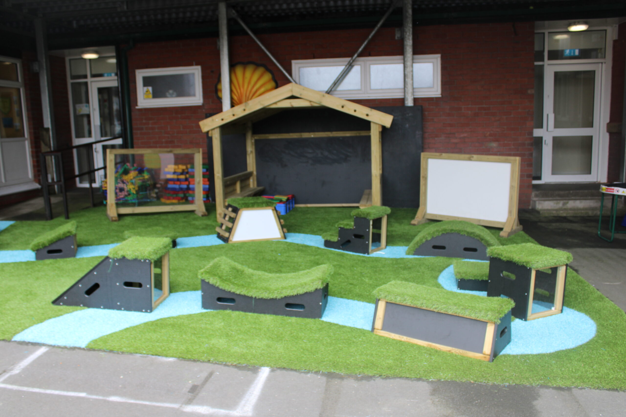 Playground area with artificial grass, blue pathways, modular play structures, a small wooden shelter, and two Mark Making Drywipe Boards - Moveable in front of a brick building.