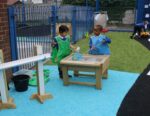 Two young children wearing aprons play with water and sand at an outdoor water table in a fenced playground area.