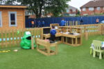 Children play in a fenced outdoor area with wooden play kitchen sets, a dedicated Mud Kitchen Corner, artificial grass, and a green frog sculpture near a wooden playhouse; houses are visible in the background.