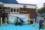 Three young children in school uniforms play with a large Mark Making Water Wall, using tubes and ramps to guide water on a blue rubber surface beside a brick building.