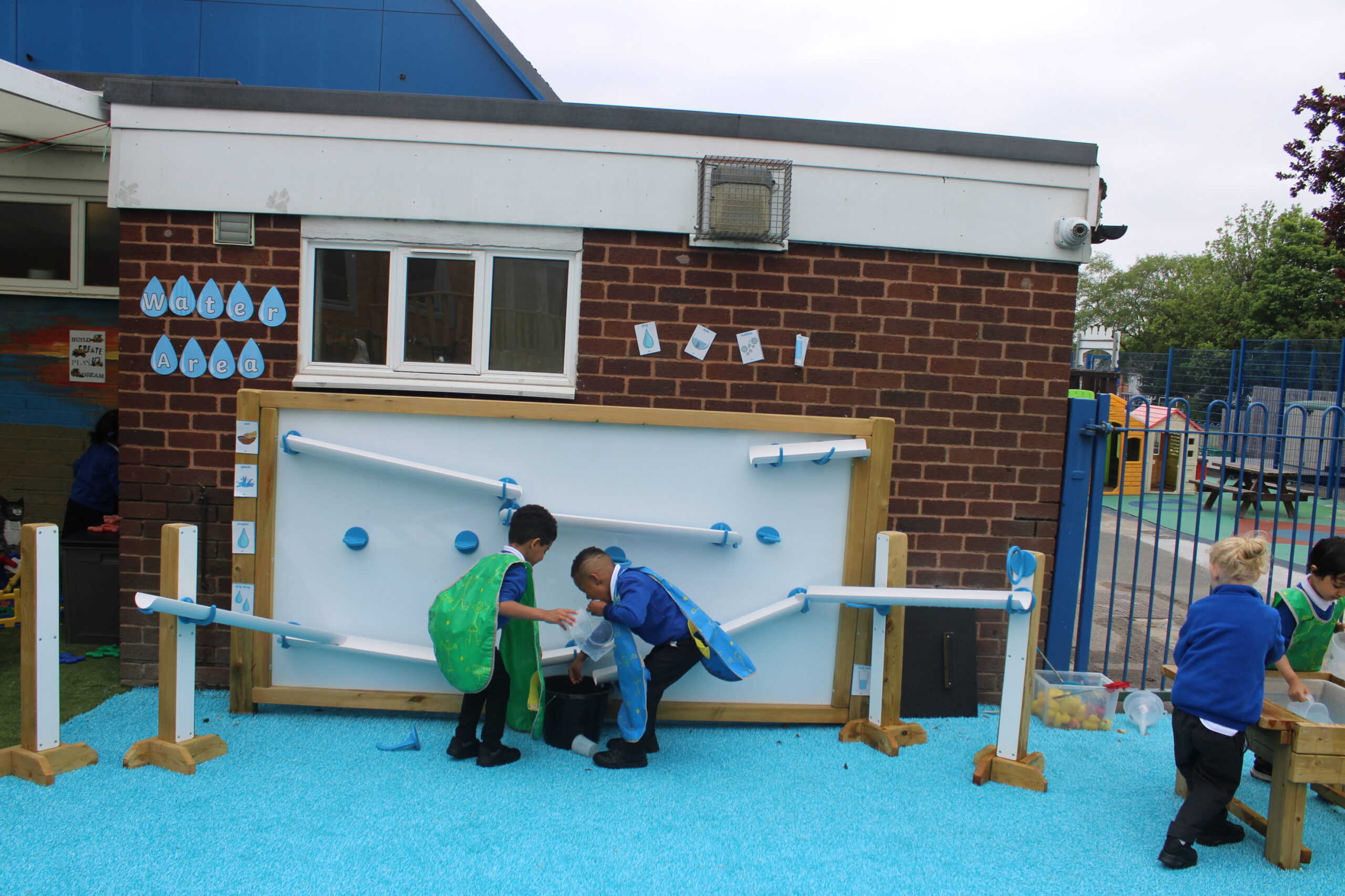 Three young children in school uniforms play with a large Mark Making Water Wall, using tubes and ramps to guide water on a blue rubber surface beside a brick building.