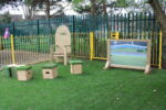 A wooden chair, four cube stools with artificial grass tops, and a large Mark Making Mirror are arranged on grass near a fenced playground area.