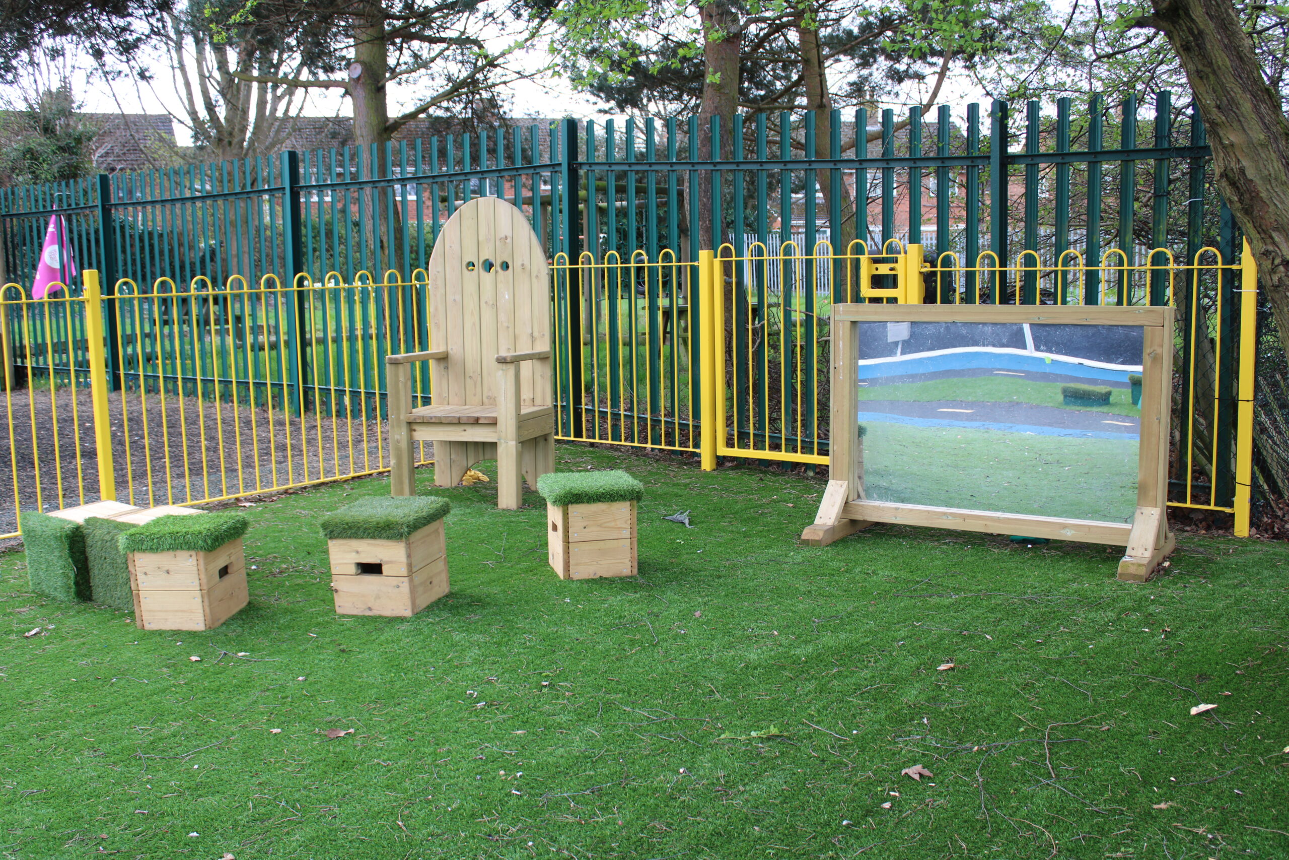 A wooden chair, four cube stools with artificial grass tops, and a large Mark Making Mirror are arranged on grass near a fenced playground area.