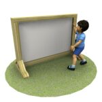 A young child in blue clothes stands on grass and touches a large wooden-framed whiteboard or display board.