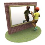 A child in a red cap and yellow shirt looks at their reflection in a large mirror set in a brick wall on a patch of grass.