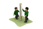 Two children in green jumpers touch opposite sides of a vertical wooden pole with metal bars, standing on a patch of green ground.