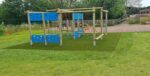 A wooden playground climbing structure with blue panels, inspired by the Enterprise Space Frame, stands on artificial grass in a grassy outdoor area, with trees and fencing in the background.