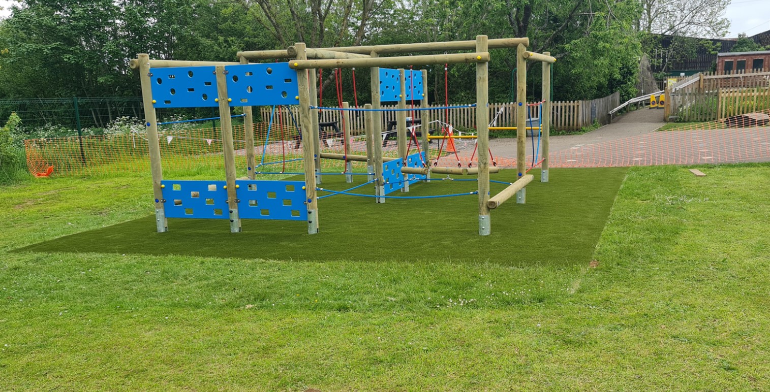 A wooden playground climbing structure with blue panels, inspired by the Enterprise Space Frame, stands on artificial grass in a grassy outdoor area, with trees and fencing in the background.