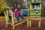 Four children play on a wooden playground structure with ropes and a tower, surrounded by trees and autumn leaves.