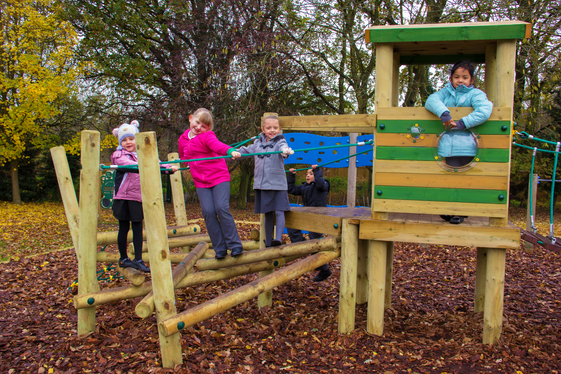 Four children play on a wooden playground structure with ropes and a tower, surrounded by trees and autumn leaves.