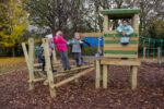 Several children are playing on a wooden climbing frame and playhouse in a playground surrounded by trees and fallen leaves.