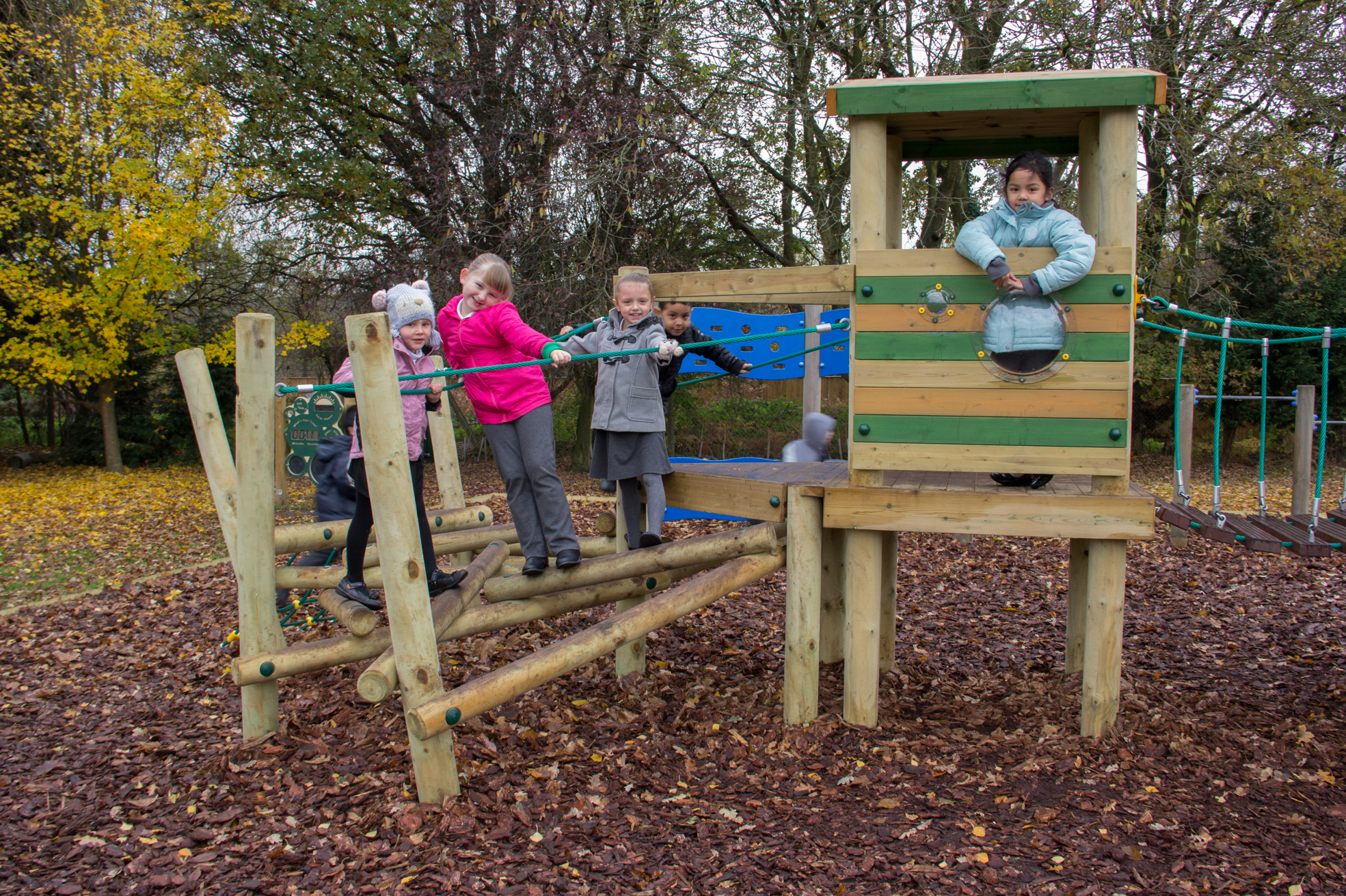 Several children are playing on a wooden climbing frame and playhouse in a playground surrounded by trees and fallen leaves.