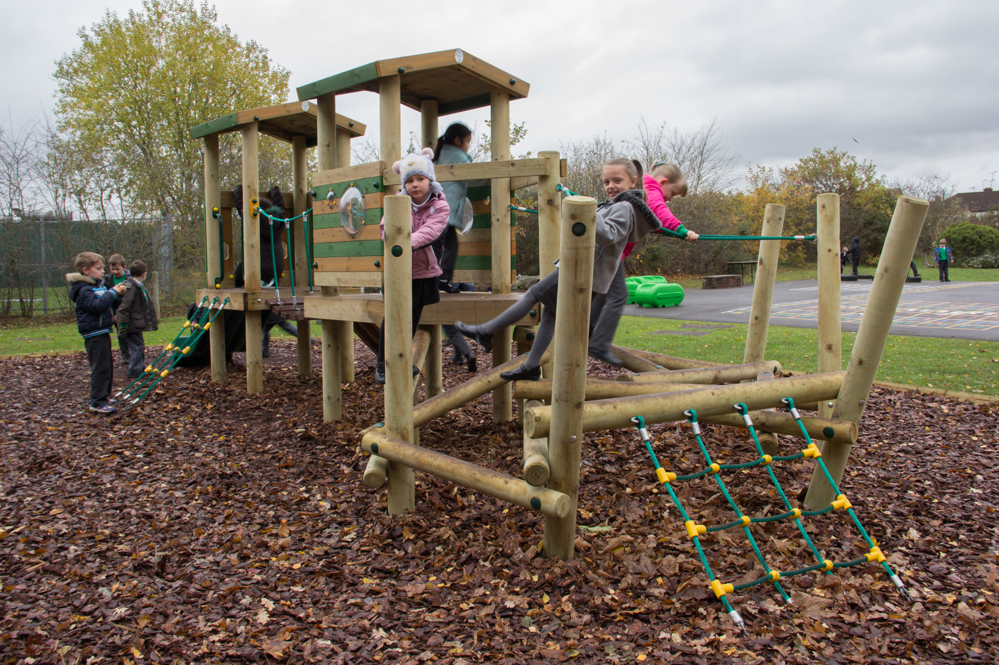 Children play on a wooden playground structure surrounded by fallen leaves on a cloudy day. Some climb ropes whilst others stand or walk on the equipment.