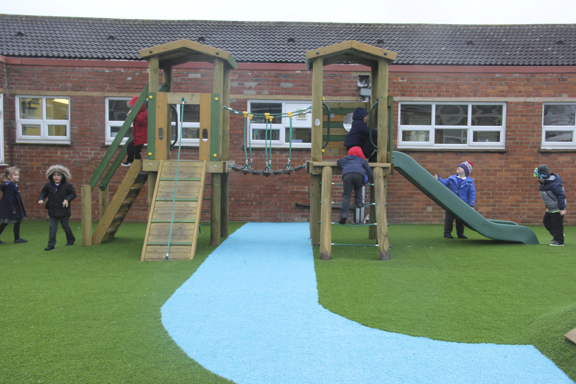 Children play on a wooden playground structure with slides and climbing ramps, set on artificial grass outside a brick building.