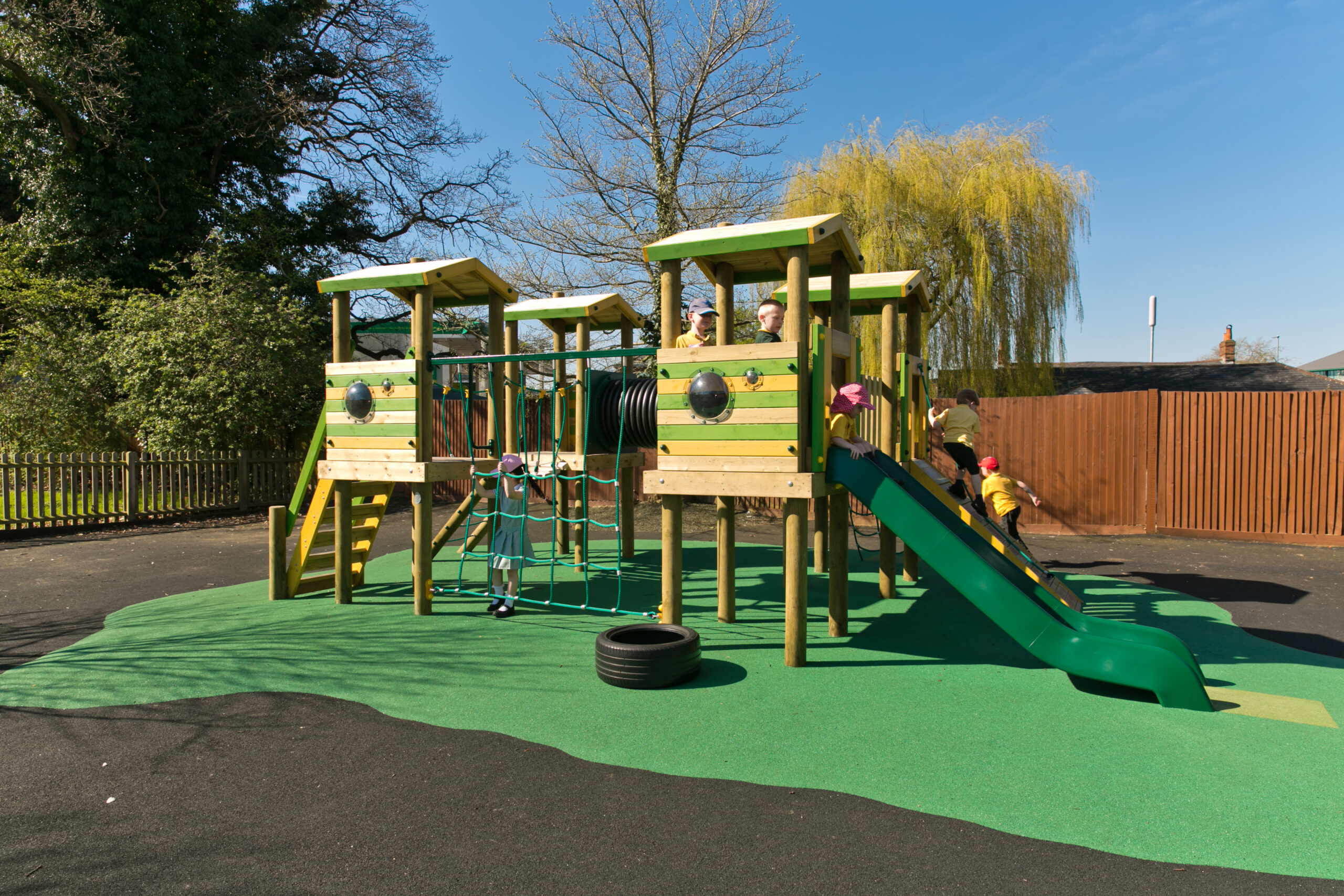 Children play on a green and wooden playground structure with slides, climbing nets, and platforms, set on a green safety surface under a clear blue sky.