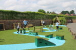 Children play on artificial grass with blue painted paths and various wooden and turf play structures in an outdoor playground area.