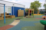 A school playground with colourful play equipment, including a Rainbow Arch and wooden structures. Children stand near a frame with coloured panels, and the ground is covered with rubber safety flooring.