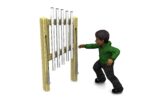A child in a green hoodie stands next to an outdoor musical chime instrument, reaching towards the metal tubes with a mallet in hand.