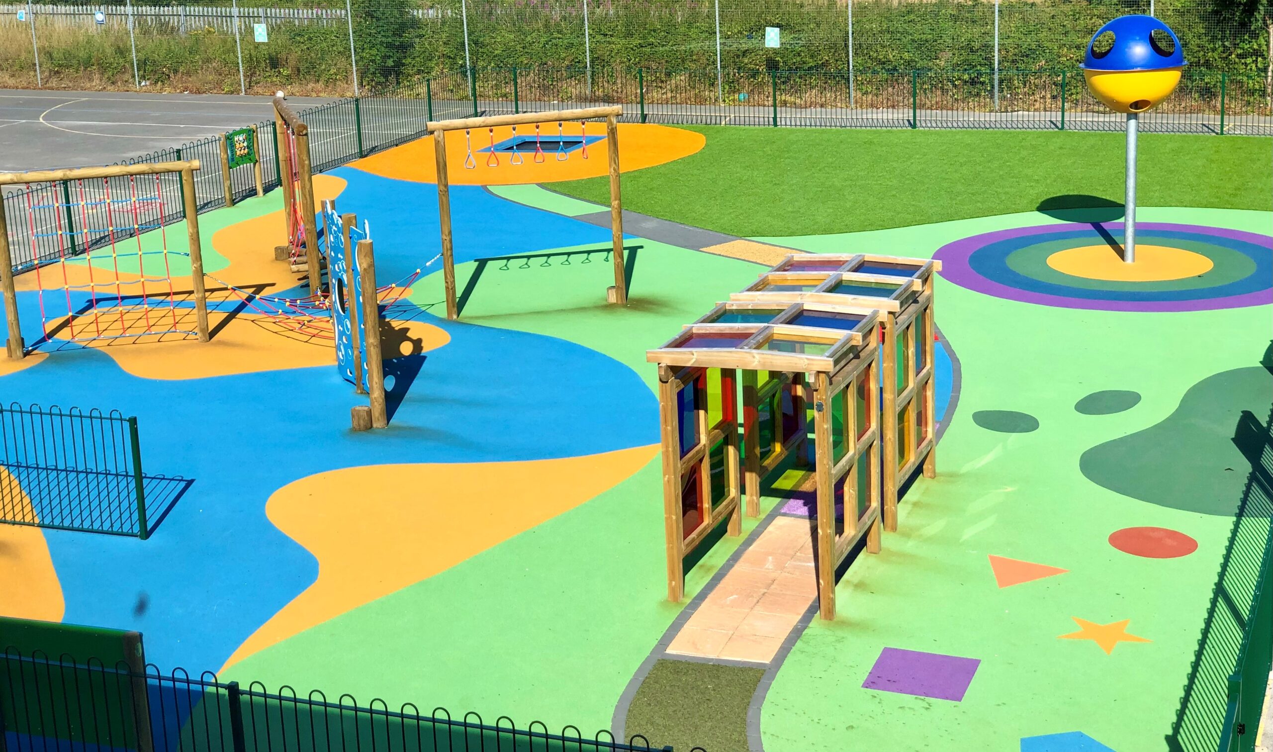 Colourful playground with climbing frames, rope bridges, and a spinning globe structure on a patterned rubber surface, surrounded by a fenced area.