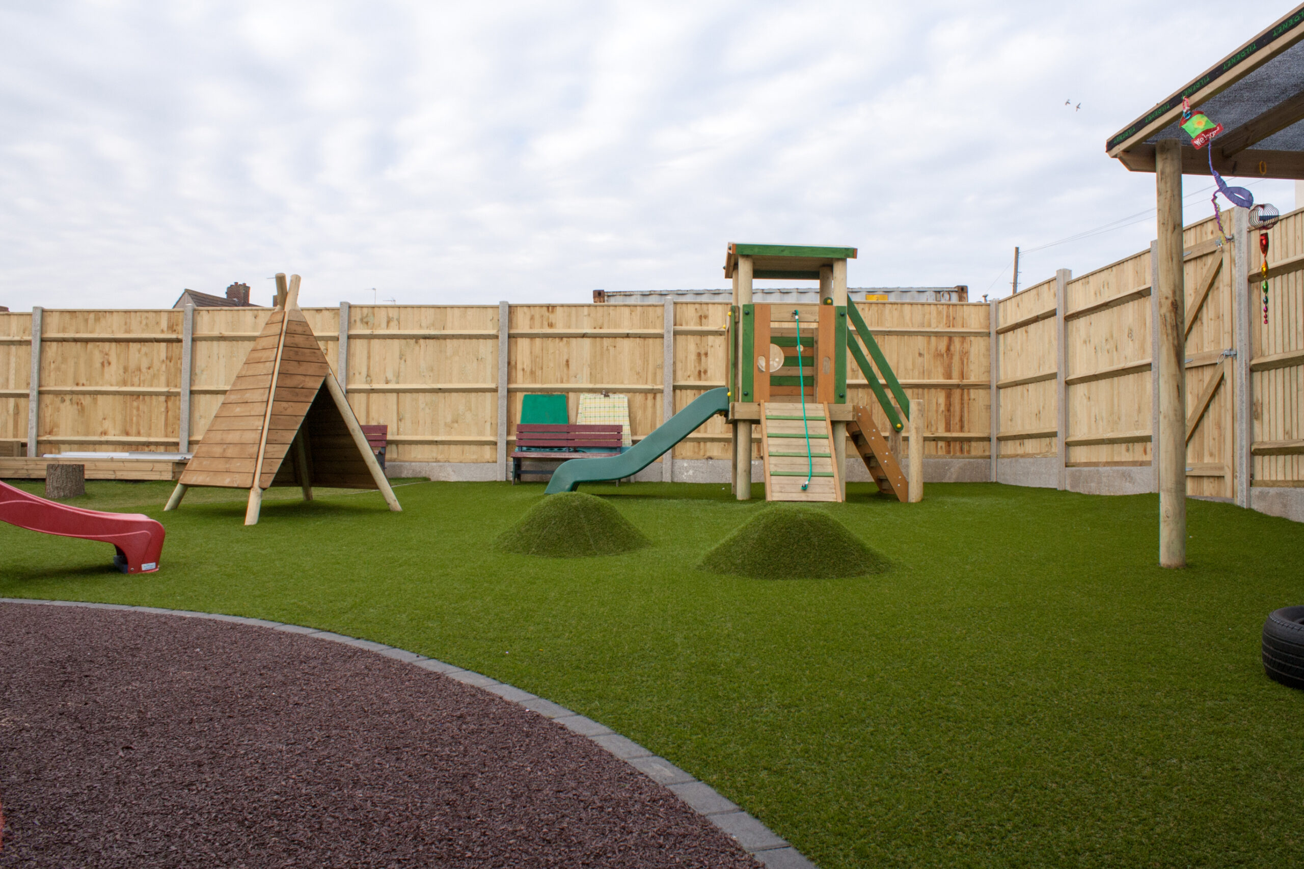Outdoor playground with artificial grass, slide, climbing structures, bench, and wooden fence surrounding the area under a cloudy sky.