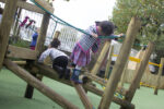 Two children climb on a wooden play structure in an outdoor playground with green flooring. Other children and adults are visible in the background.