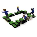 Four children in blue shirts play on a modular indoor obstacle course made of green and black blocks, steps, and ramps.