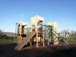 Wooden playground structure reminiscent of Fort Knox, with slides, climbing nets, and ladders on a grassy area under a clear blue sky. Trees and hills are visible in the background.
