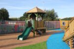 Three children in school uniforms play on a Takamaka 1200mm with Poly Slide, enjoying the wooden playground structure within a fenced area and colourful safety surfacing.