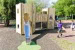 Children play near the Fort Cumberland with Steel Slide, a wooden playground structure featuring climbing walls and rope elements on a sunny day.