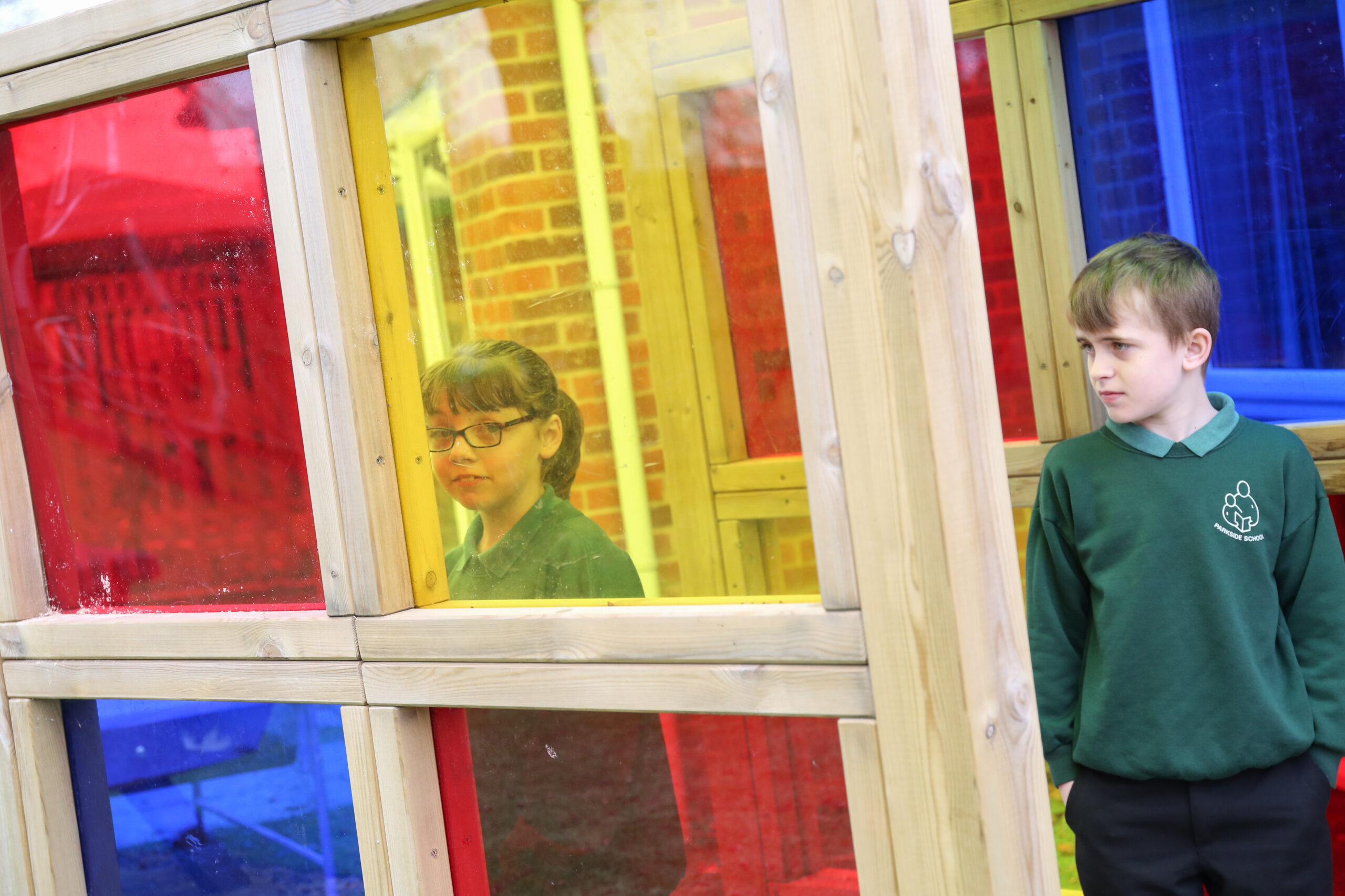 Two children in green uniforms stand near multicoloured glass panels; one child is behind the panels, and the other stands outside looking towards the panels.