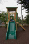 A young child sits at the top of a green slide attached to a wooden playground structure, preparing to slide down. Trees and a fence are visible in the background.