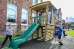 Children play on a wooden playground structure with a slide and climbing bars outside a brick school building on a sunny day.