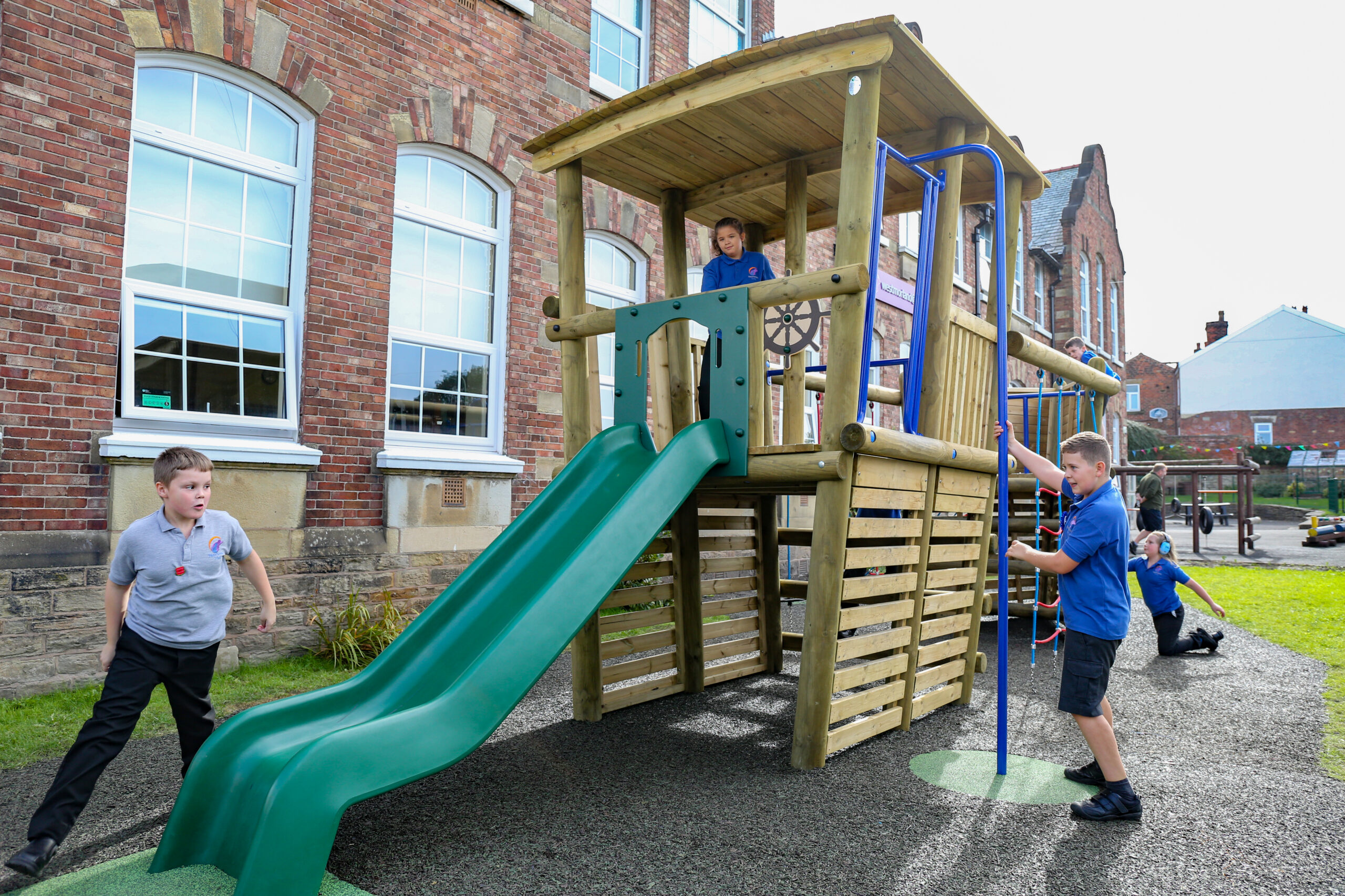 Children play on a wooden playground structure with a slide and climbing bars outside a brick school building on a sunny day.