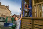 Two children play on a wooden playground structure; one stands on the ground holding a blue pole whilst the other is on the platform above, smiling.