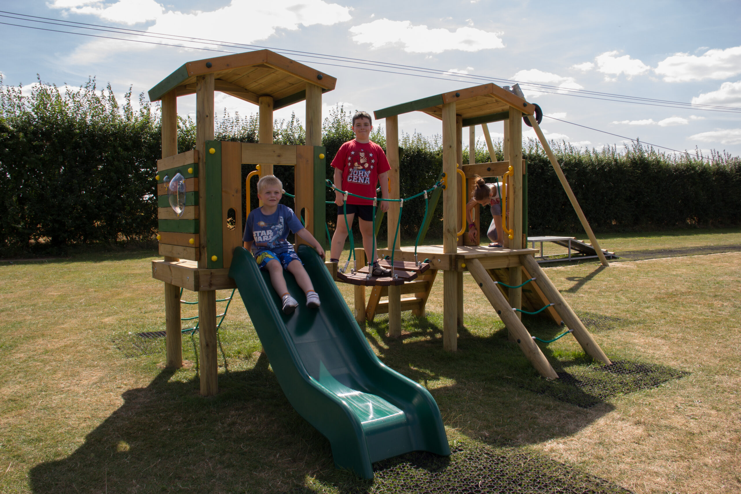 Two children play on a Young Explorer - Marco Polo 1200-1500mm wooden playground set with a slide and rope bridge, while an adult stands nearby on a grassy field under a partly cloudy sky.