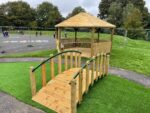 A small wooden footbridge leads to a gazebo on artificial grass in an outdoor area, with a fenced playground and people in the background.