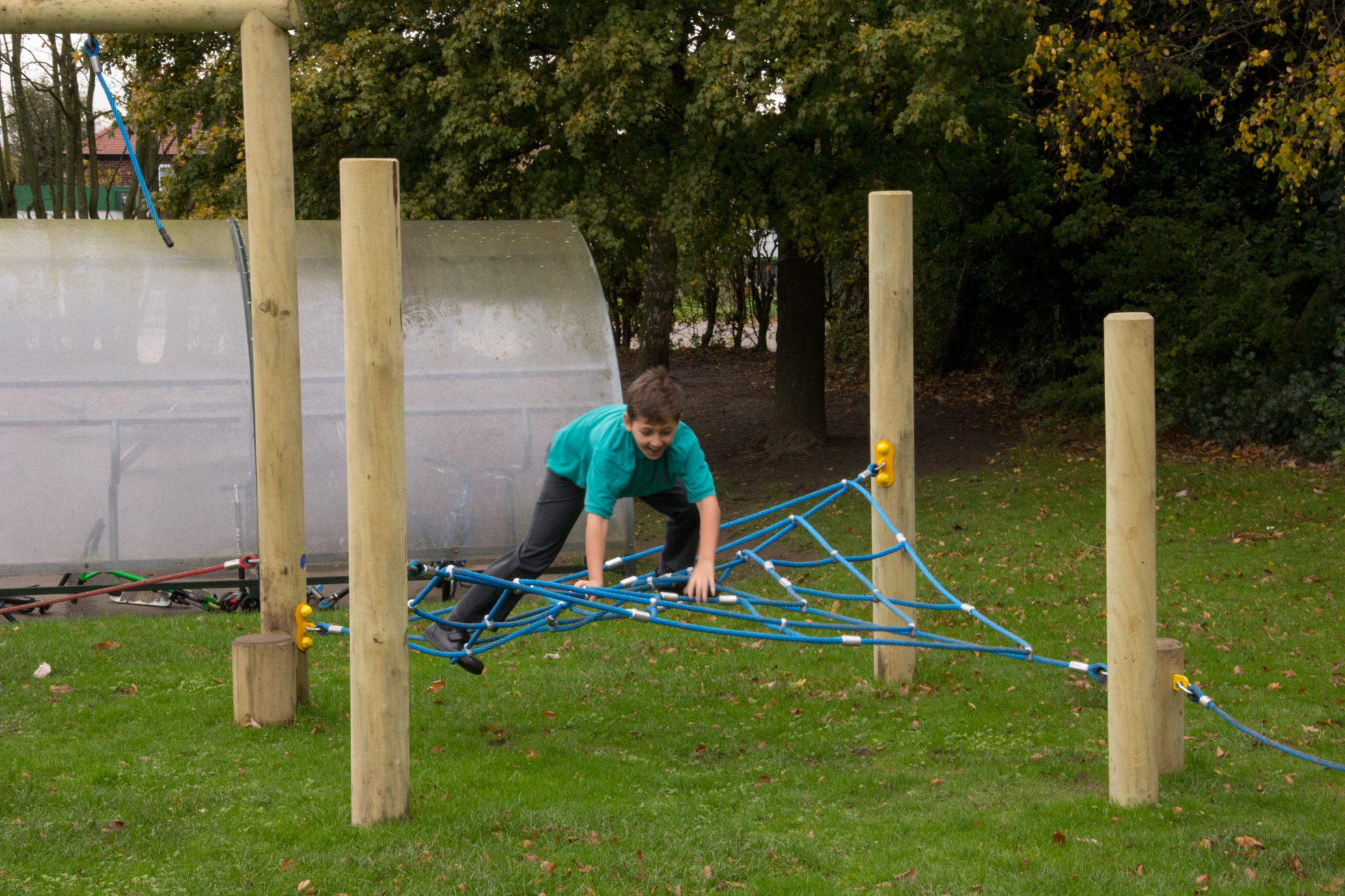 A boy in a green shirt climbs on a blue rope structure in an outdoor play area surrounded by grass and wooden posts.
